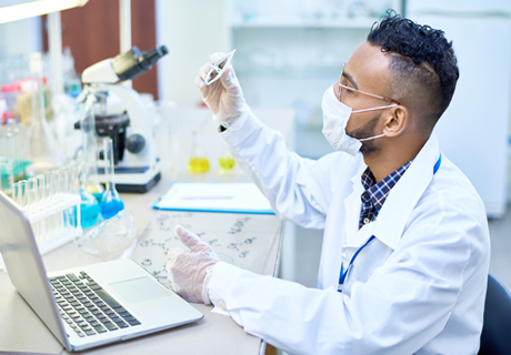 A male lab technician viewing a specimen and recording notes on a laptop